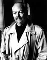 black and white photo of a middle-aged White man who looks intelligent, sensitive and refined; he has a neatly-trimmed mustache and is wearing a white trenchcoat over a black turtleneck.