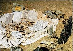 photo of debris in the dirt - book pages and torn bits of paper lying on a white cloth, perhaps a doorknob, various other unidentified objects.