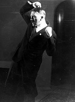 Old photo of Hitler standing up, leaning backward at an odd angle with his clenched fists in the air, one fist above his head and the other by his shoulder.