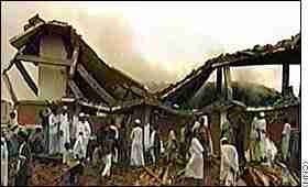 Sudanese men dressed in white robes and white caps, standing around the smoking ruins of their medicine factory, destroyed by U.S. cruise missile
