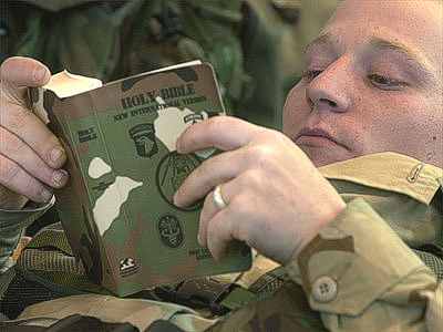 photo of a self-satisfied looking soldier wearing a camouflaged army uniform as he reads a similarly camouflaged Holy Bible.