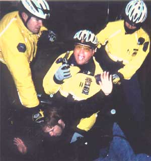 Photo of three police-thugs wearing florescent yellow jackets at night, holding a young White man down on the ground. The young man has his arms spread wide, hands up in a gesture of surrender, yet the pig on the left is gratuitously gripping the young man's hair.  The pig in the middle is holding a handgun and pointing it up in the air slightly toward the viewer. His big mouth is open wide as he yells something.