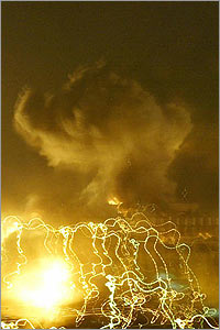 photo of nighttime explosion, bright yellow-white flash in the foreground with a web of yellow-white tracer lines around it, and a large mushroom cloud of white smoke in the background, fires burning below it.