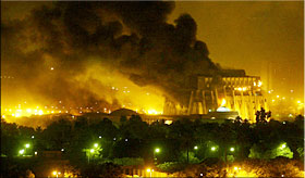 nighttime photo shows a large building in the distance with a huge plume of black smoke rising out of it; behind it are many fires burning bright yellow-white.