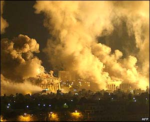 nighttime photo of many buildings engulfed in white smoke, smoke rising in tall plumes into the night sky; what looks like an explosion of white smoke just occuring on the left side; all the smoke lit up by the light of fires to the right.