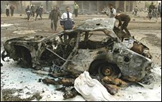 photo of a totally burned, carbonized car on a street, with many people in the distance behind it; there is white ash or some powdery substance all around the car and the paint and tires are completely burned off; next to the car is what looks like a burned body, with perhaps another body under a white cloth next to it.