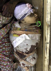 Photo of a dead baby in a wooden coffin. He is wrapped in cloth, dried blood soaking through the cloth, a lavender cloth wrapped around his head with a little flower attached.