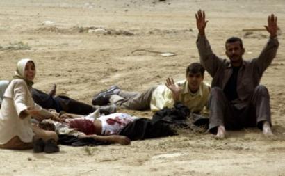 photo of a group of five people sitting or lying on the grayish-brown sand; at least one person is bleeding from wounds, red blood staining his white shirt and pants as he lies on his back; near him is a distraught woman with her hand on his shoulder, her other hand held out beseechingly toward the viewer, an anguished look on her face; to the right a man sits in the sand facing the viewer with his hands held above his head, looking pained and worried; behind him a teenage boy lies on his stomach with one hand held up, looking fearful; another body is lying on the sand behind the woman but only the legs are visible.