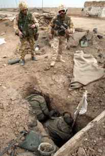 Photo of two dead men huddled in a rectangular trench in the dirt, a white flag on a pole beside them, leaning against the wall of the trench.  Above them, looking down at them, stand two heavily armed soldiers.
