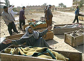 Photo of a man wearing a black-and-white Arab headdress and a dark gray robe, kneeling beside one of at least seven wooden coffins which are lying in the middle of a dirt street. The man is crying as he spreads his arms wide and closes his eyes as he raises his face up toward the sky.  Other men are standing and walking around behind him, among the other coffins.