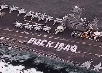 Photo of the deck of an aircraft carrier, taken from one or two hundred feet above as it plows through the ocean water. Many white aircraft with folded wings line the deck.  A large group of sailors and/or pilots wearing white uniforms are standing on the angled runway of the deck, having formed themselves into two large white words which read: FUCK IRAQ.