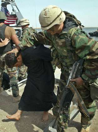 Photo of a helmeted soldier in green fatigues, carrying a machine-gun with strip of bullets hanging out of the side. The soldier is holding his hand on the back of the head of a young barefoot Iraqi man, forcing him to bend down forward with his face parallel to the ground as he walks with his hands tied behind his back.