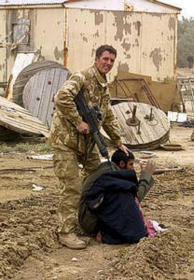 Photo of an evilly leering American soldier looking into the camera as he pokes the barrel of his rifle into the shoulder of an Iraqi man sitting in the dirt below him. The soldier is holding his other hand on the top of the Iraqi man's head, pushing it down while the Iraqi man has his hands in front of him in a gesture of prayer.