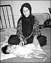 A gaunt-looking Iraqi mother is sitting on a bed looking down at her emaciated little child who is lying on the bed and gazing with big dark eyes at the viewer.