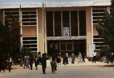 A large tan and white building, looking slightly run down, with a sign in Arabic over the entrance, and evergreen trees on either side of the entrance. Various students are standing around and walking in the open area in front of the building.