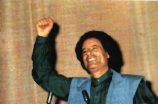 A young Qadhafi smiling and raising his right fist in the air as he stands before microphones, apparently in front of a crowd of well-wishers.  He is wearing a dark green shirt with a light blue vest.