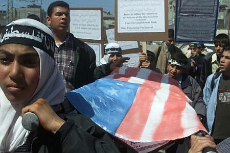 photo of a crowd of Palestinian boys of varying ages carrying signs in English and Arabic, with four boys bearing a symbolic stretcher over which is draped a homemade American flag. The ceremonial stretcher bearers are wearing white Arab headdresses with wide black headbands on which something is written in white Arabic lettering. They all look very sad and angry.