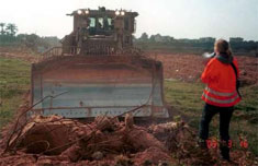 photo taken from behind Rachel as she stands before the bulldozer, wearing her bright red jacket and holding her megaphone. The bulldozer is farther away in this photo, facing her with it's shiny steel blade.