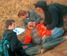 Three friends of Rachel crowd around her as she lies semi-conscious in the dirt, still wearing her bright orange-red jacket. Her head looks so pale and fragile, her eyes are closed and her mouth open, with blood on the left side of her face. There is dirt all over her clothes and her legs are bent at the knees. Two young men kneel in the dirt on either side of her as they stare at her in shock, one with his hand on her shoulder, while a woman holds Rachel's head in her hands.