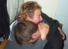 A woman cries bitterly with her eyes closed as she hugs a young man who droops his head over her shoulder.  These are two of the three friends who knelt around Rachel in the photo above, right after she had been crushed.