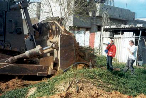A monstrous steel bulldozer on the left, so big you can only see the front of it and the blade. To the right, in front of the huge blade is Rachel Corrie, looking very small by comparison. The blade alone is so big it's several feet higher than her head. The rest of the bulldozer rises above that. Rachel is wearing a bright red florescent jacket with white florescent stripes, and holding a megaphone as she talks to the driver. Standing next to her is a young White man, one of her fellow activists. Behind them is some ramshackle corrugated metal fencing and a plain, gray, two-storey concrete house.