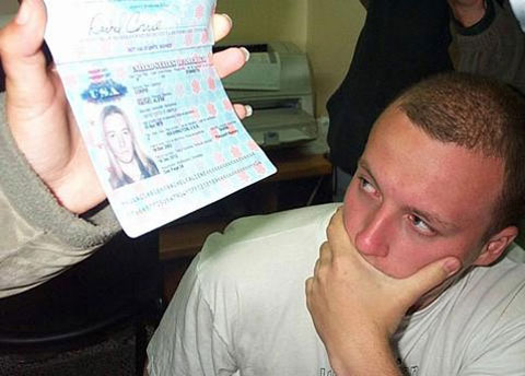 A young White man with a crewcut, wearing a white t-shirt, looks at Rachel's passport and photo as somebody's hand holds it up for the camera. The young man's face is red and he holds his hand over his mouth as he looks at the photo.