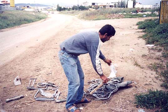 Photo of a Palestinian man bending over the broken and flattened remains of a wheelchair by the side of a dirt road.  He is picking up the small white flag attached to a wooden stick that was on the wheelchair. It looks like there is dried blood soaked into the dirt underneath the crushed wheelchair.
