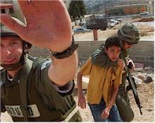 Photo shows a helmeted Israeli soldier with a pudgy, boyish, pink face who smiles and holds his hand up to block the camera. Behind him another helmeted Israeli soldier carrying a rifle has his arm in a choke-hold around the neck of a Palestinian boy of about 12 years of age, forcing him to walk somewhere to the right of the viewer.