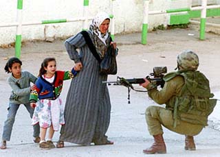 photo of an Israeli soldier squatting on the ground as he points his rifle at two little girls who are hiding behind their mother. The mother is crying as she grips one of her little girls' hands and looks at the soldier, and the little girls have expressions of fright on their faces as they look at the soldier.
