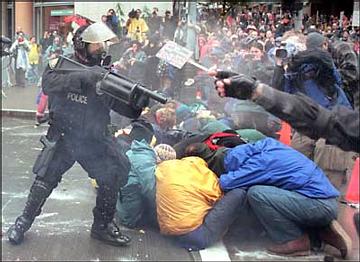 Photo of an armored, black-clad pig pointing a black teargas-shotgun at a crowd of people, some of whom are sitting on the street as they lean forward and cover their faces protectively.  A white mist of tear gas is in the air around the pig and above the heads of the people.