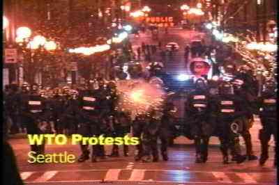 photo of TV screen image shows a phalanx of black-clad pigs on a street in downtown Seattle at night, advancing toward the viewer - a yellow burst of sparks in the midst of the phalanx indicates one of the pigs is firing tear gas toward the viewer.  Words at bottom of screen read 'WTO protests - Seattle'.