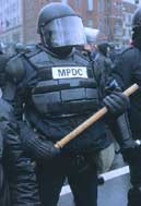 Photo of a black-clad, heavily-armored robo-pig, visor hiding his face, holding his hardwood police baton in both hands prominently in front of him.