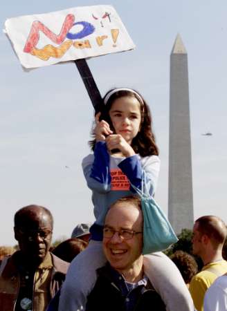 photo of a little girl riding on her father's shoulders as she holds a sign which says 'No War!'. The Washington Monument is in the background.