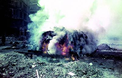 photo shows a blackened pile of burning bodies, white smoke billowing up, amidst the rubble of the city.