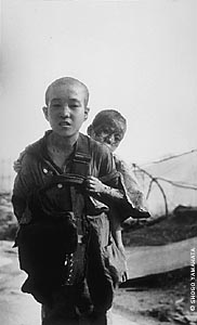 Photo of a Japanese boy carrying little child on his back. The child is peeking over the shoulder of the older boy at the viewer, and the skin on little child's forehead and cheeks and around his eyes looks blackened and burned.