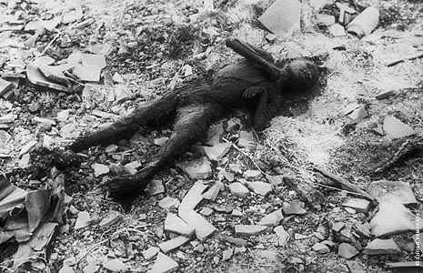 Photo of the blackened corpse of a small child lying on his back on the ground. The ground is grayish-white, covered with little rocks and what looks like pieces of broken pottery.