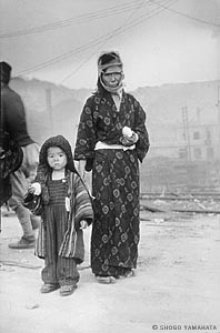 Photo of a Japanese woman wearing a kimono, standing next to a little child of around three. The air around them looks dusty, and the background in the distance is obscured by haze. The child is wearing a coat with a hood, and he looks at the viewer with a curious and worried expression on his little face.