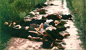 A group of dead Vietnamese women and children lying on a narrow dirt road, with tall green grass on either side of it.  A woman in the foreground appears to have a small pool of her blood in the dirt around her head; there are naked dead babies sprawled here and there and several small dead children.