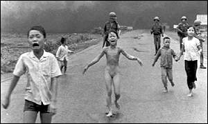 Black and white photo of five Vietnamese children running in pain and fright down a road toward the viewer.  Behind them are four soldiers, either South Vietnamese or American, and in the distance is a huge wall of gray smoke from the napalm attack.  The boy closest to the viewer is crying in open-mouthed terror; behind him in the center of the photo is nine-year-old Kim Phuc screaming in pain as she runs, her clothes burned off by the napalm; behind her to the right another girl her age is running as she holds the hand of her little brother; and another little boy is behind them to the left, looking back at the soldiers as he runs.