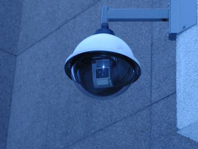 An object that looks at first glance like a street light hanging from a metal arm which is attached to the outside wall of a building. Inside the transparent lower hemisphere is the surveillance camera.