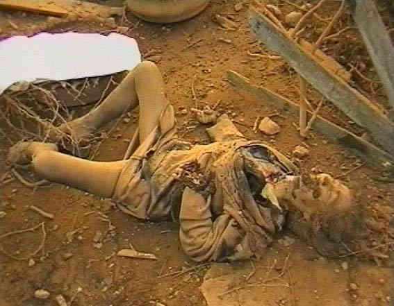 A dead woman is lying on her back in the dirt amidst debris; there is dried, dirt-encrusted blood on her mouth and face, her left hand appears to be severed and her right hand is at an odd angle; there is a bloody hole at the base of her throat; she appears to be covered with dust that makes her body and clothes the same color as the dirt around her.